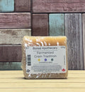 A packaged shampoo bar from Boreal Apothecary, labeled 'Fermented Grain Tradition,' wrapped in clear plastic and standing on a light wood surface against a rustic multi-colour wooden plank background.