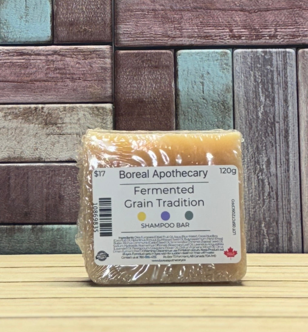 A packaged shampoo bar from Boreal Apothecary, labeled 'Fermented Grain Tradition,' wrapped in clear plastic and standing on a light wood surface against a rustic multi-colour wooden plank background.