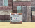 A packaged bar of Boreal Apothecary Rose Garden soap sits on a light wood surface against a rustic multi-coloured wooden plank background.