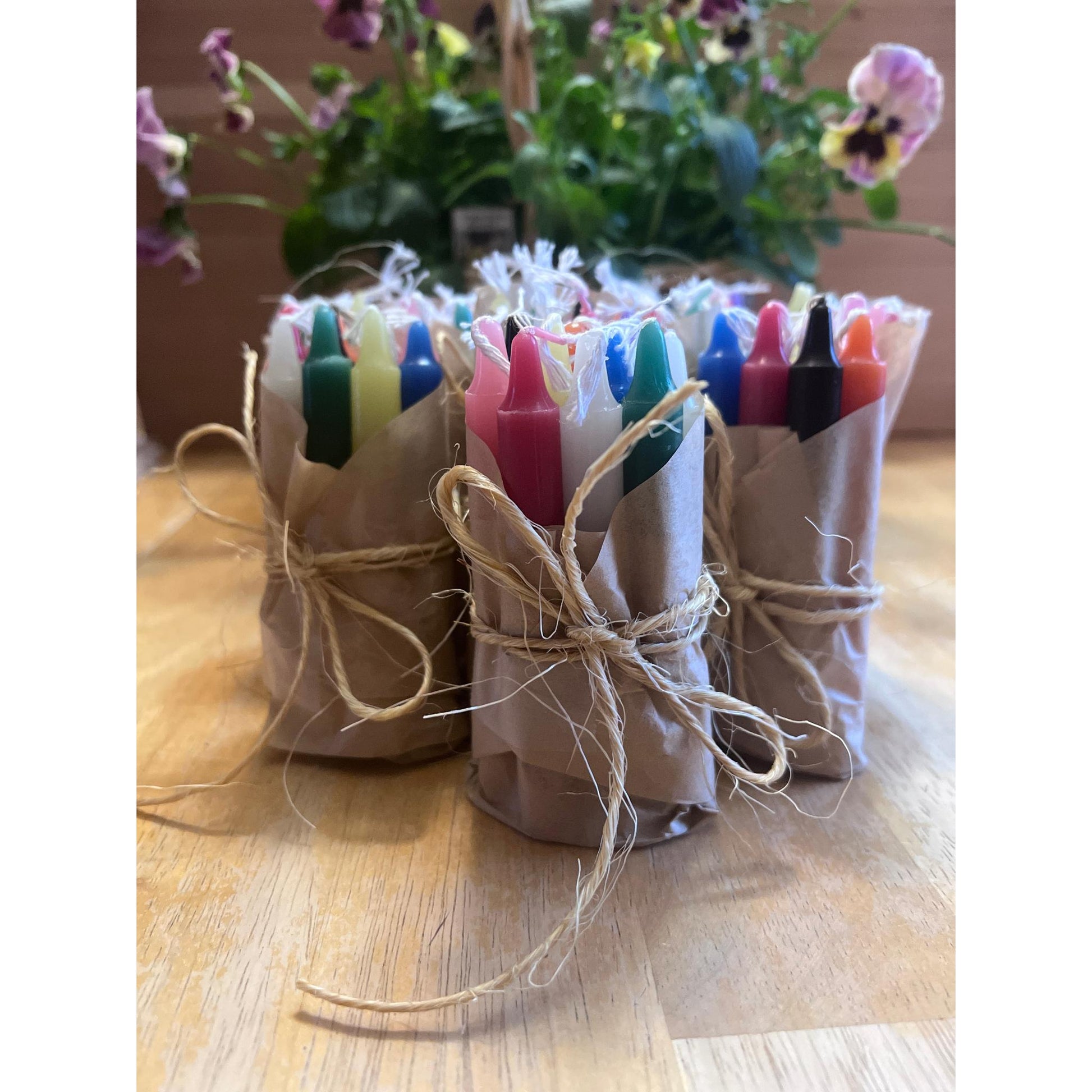 Colourful candles in paper wraps tied with twine on a wooden surface with flowers in the background.
